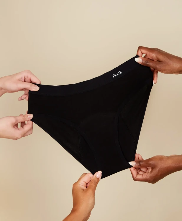 Czarne majtki menstruacyjne Fluxies Hipster na obfity okres rozciągane w dłoniach na beżowym tle