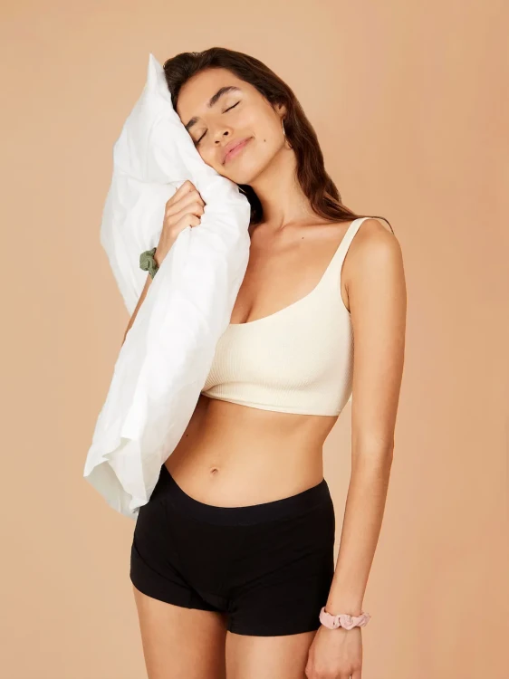 Kobieta trzymająca w ręku poduszkę w czarnych majtkach menstruacyjnych Fluxies Sleep Short na bardzo obfity okres i w białym topie